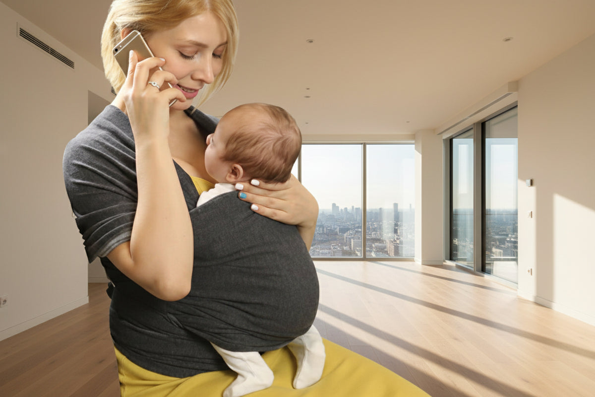 Woman holding a baby in a sling and talking on a phone in a room with furniture and bags.
