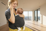 Woman holding a baby in a sling and talking on a phone in a room with furniture and bags.